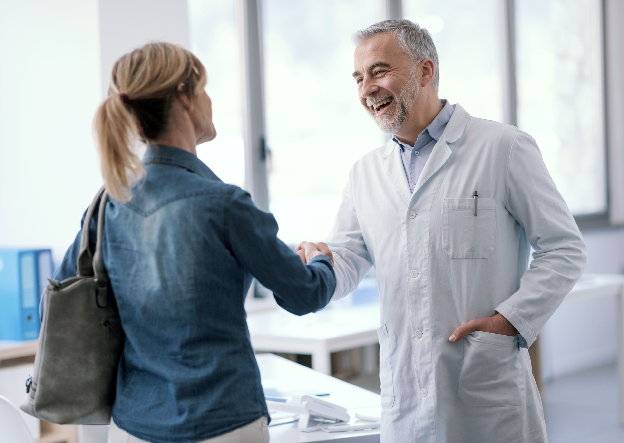 Smiling doctor in a white coat greeting a patient with a handshake in a medical office.