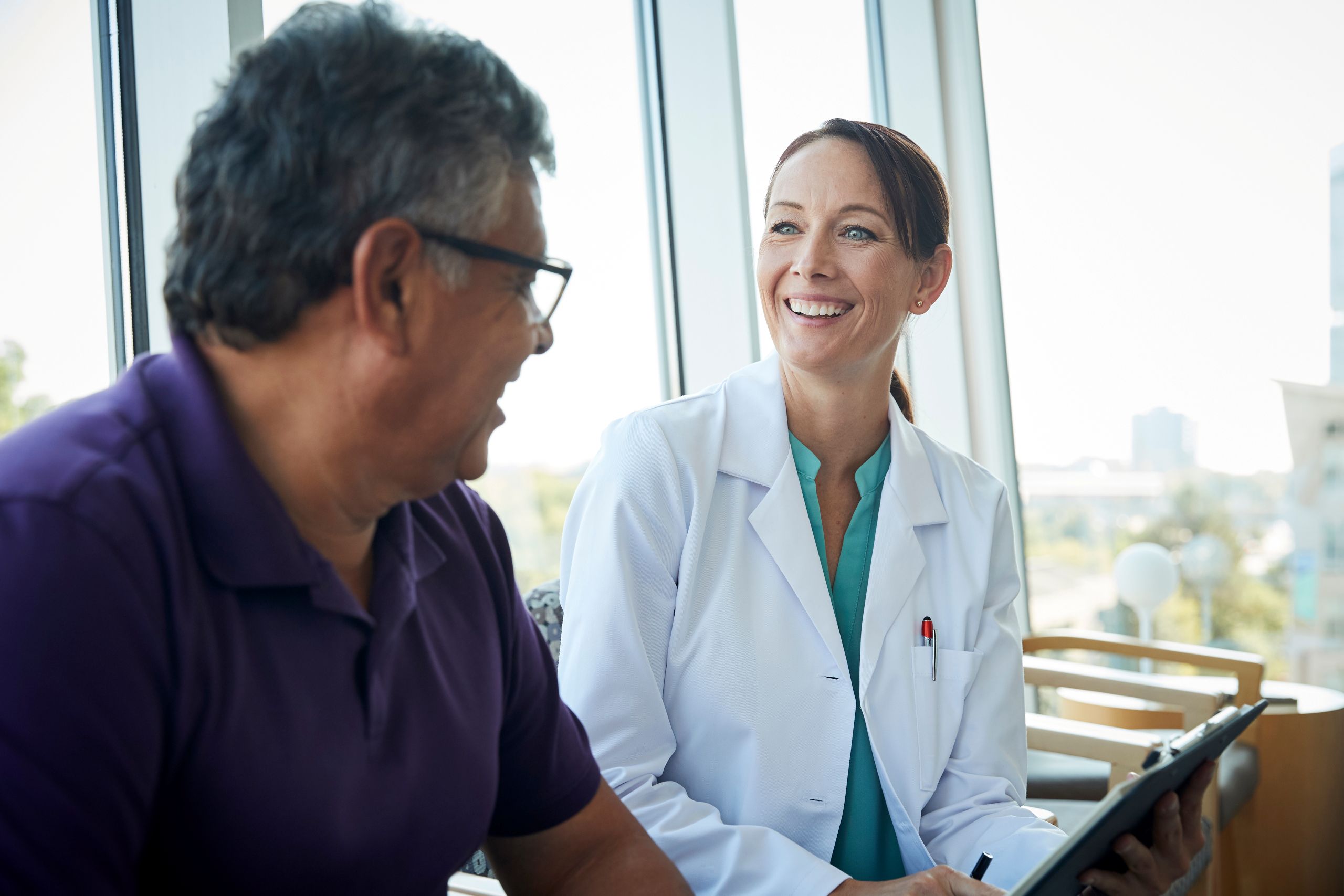 Smiling female doctor in a white coat holding a clipboard while talking with a senior male patient wearing glasses and a purple shirt in a bright medical office.