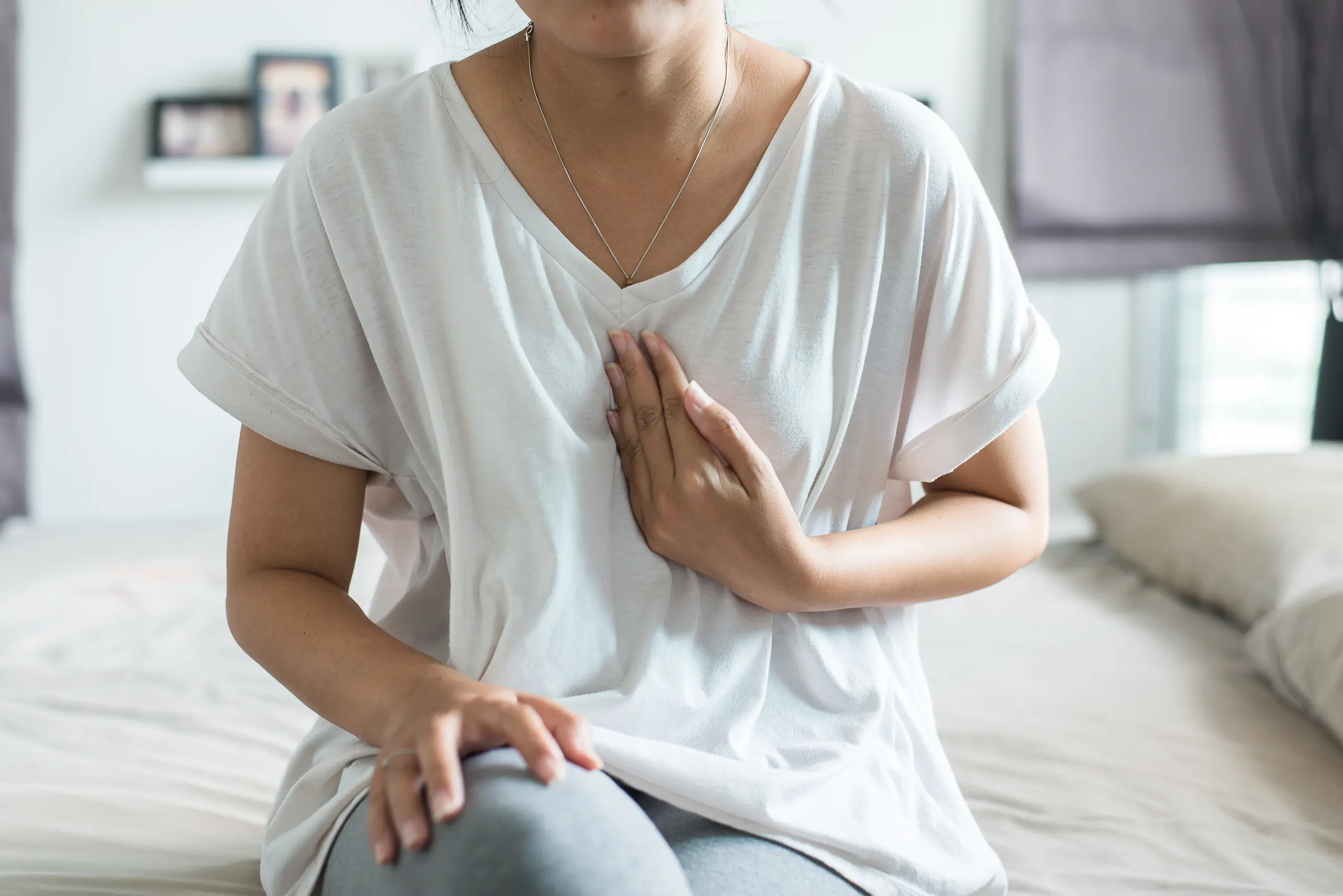 Woman sitting on a bed with her hand on her chest while experiencing heartburn or acid reflux symptoms.
